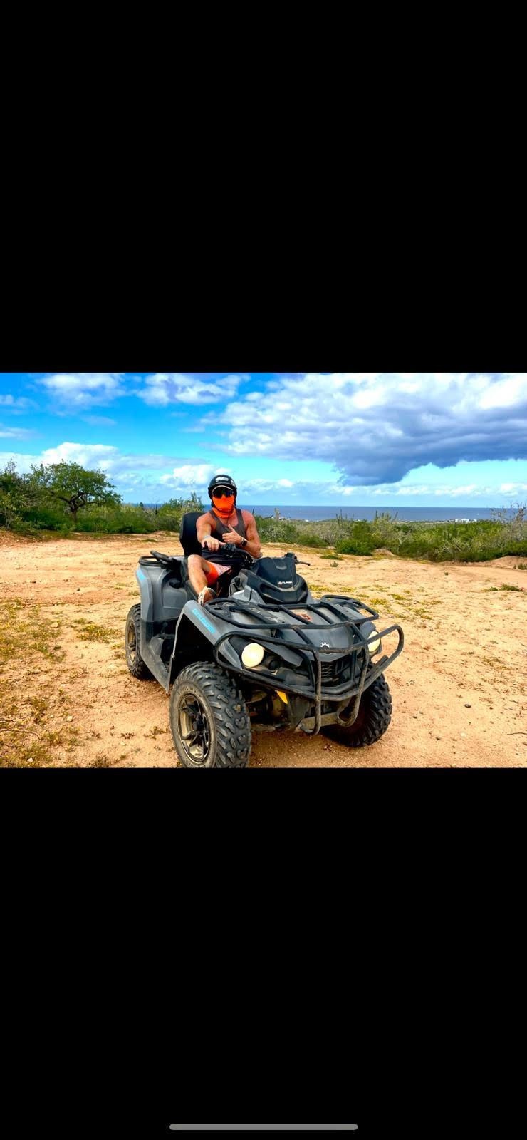 Person riding an ATV on a dirt path with green bushes and blue sky in the background.